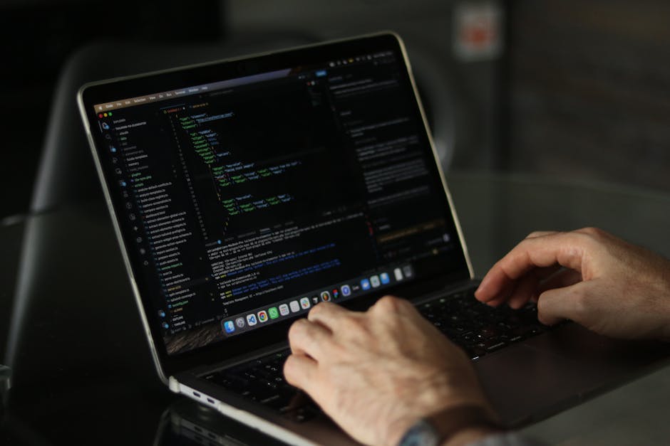 A person using a laptop with a close-up view of their hand on the keyboard, displaying lines of code on the screen in a dark-themed text editor. The laptop is placed on a wooden surface, and the indiv